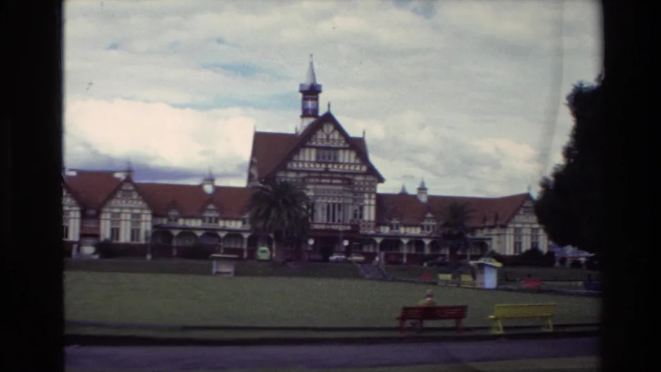 1981 New Zealand 8mm frame of a Tudor-style building viewed from a park, with benches, lawn, trees, and a distant seated person.