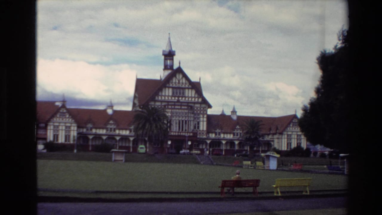 1981 New Zealand home-movie frame showing a large Tudor-style building, open lawn, benches, trees, and cloudy sky.