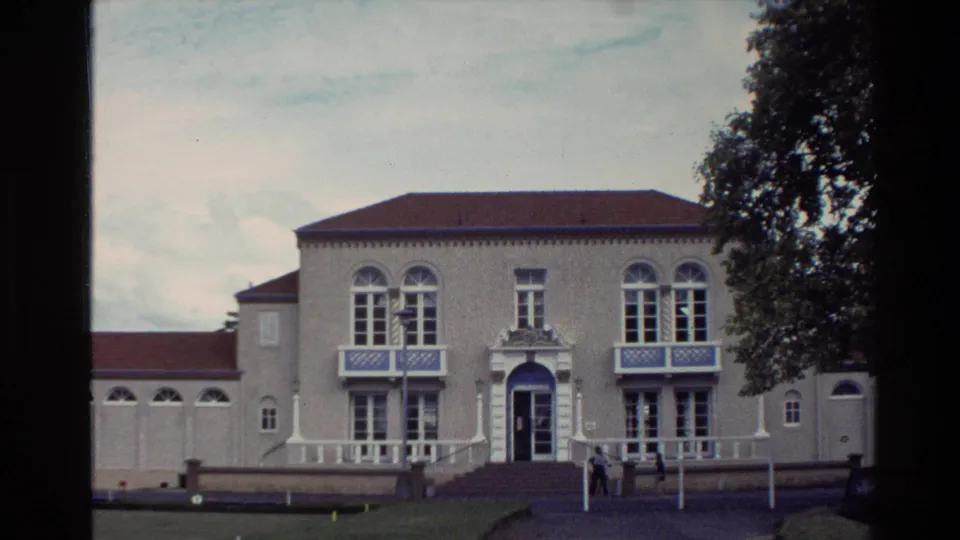1981 New Zealand home-movie frame showing the tan building front, arched windows, blue trim, lawn edge, and small figures near the doorway.