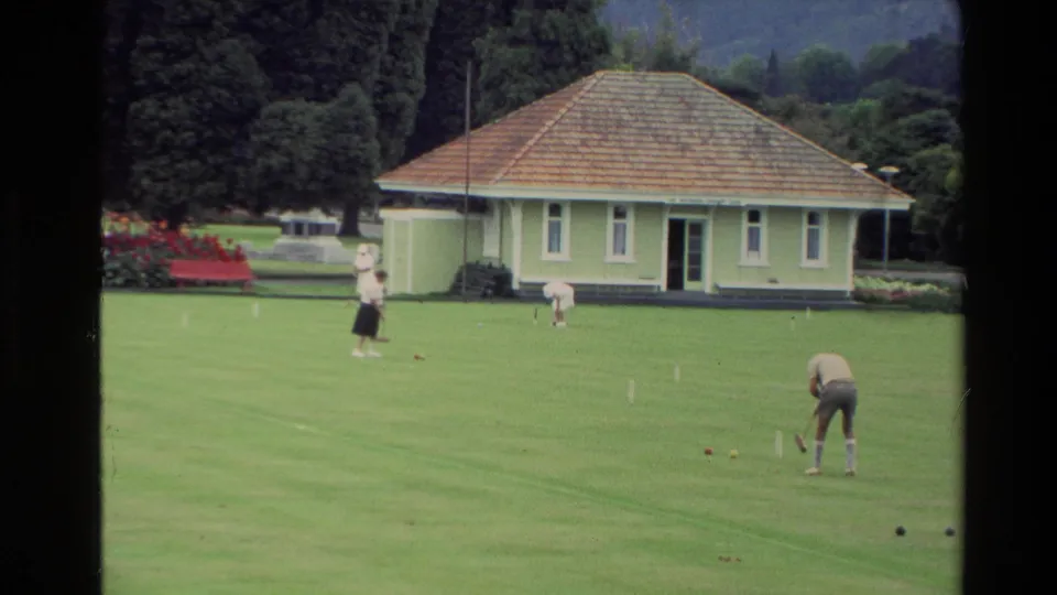 1981 New Zealand home-movie frame showing people playing croquet on a manicured lawn, with a small green pavilion, trees, and hills behind.