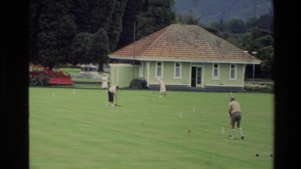 1981 New Zealand home-movie frame showing men lawn bowling on a manicured green near a small pale building, flowerbeds, and trees.