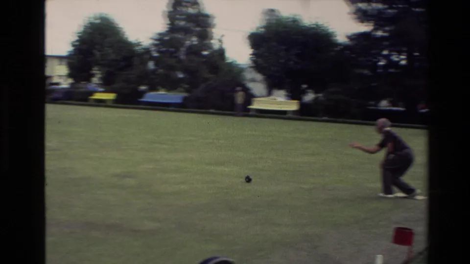 1981 New Zealand frame with a man bowling from the right side of a green, with trees, parked cars, and distant club grounds visible.