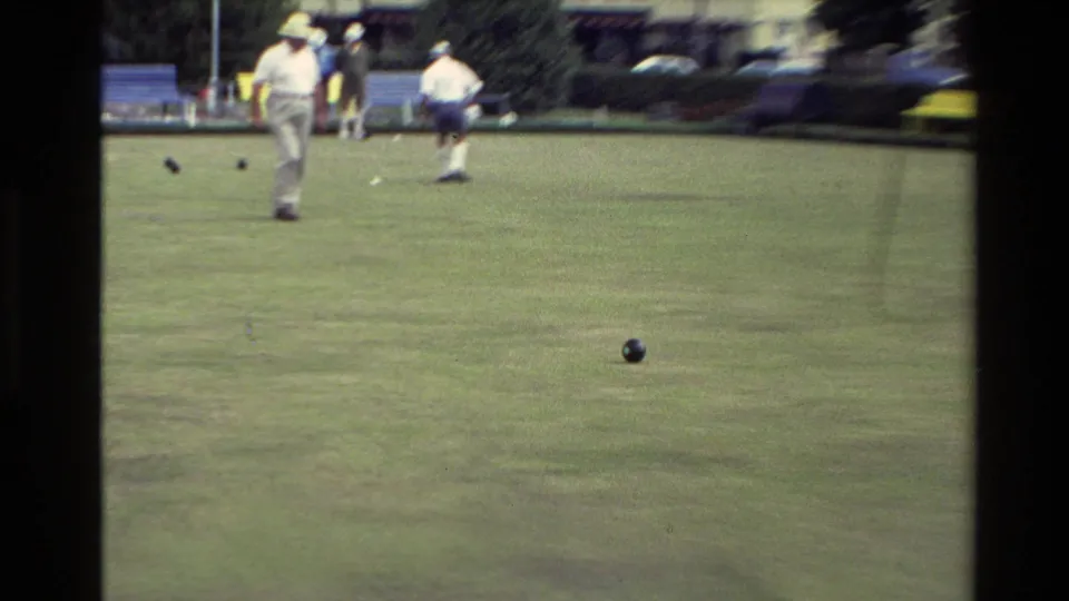 1981 New Zealand home-movie frame showing several men on a lawn bowls green, with dark bowls scattered on the grass and cars beyond.