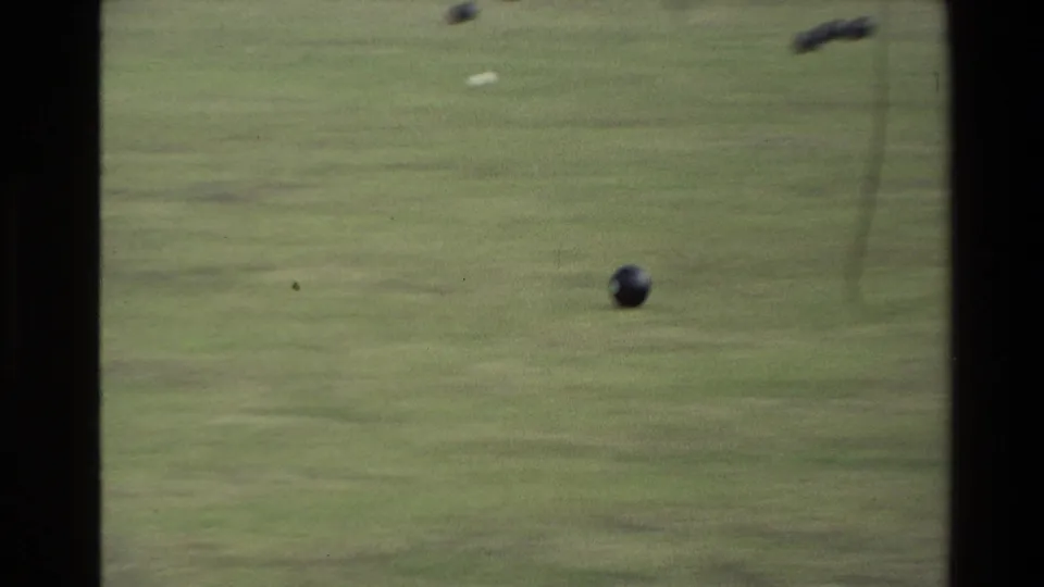1981 New Zealand frame focusing on a dark lawn bowl on the grass, with distant players and markers softened by the 8mm image.