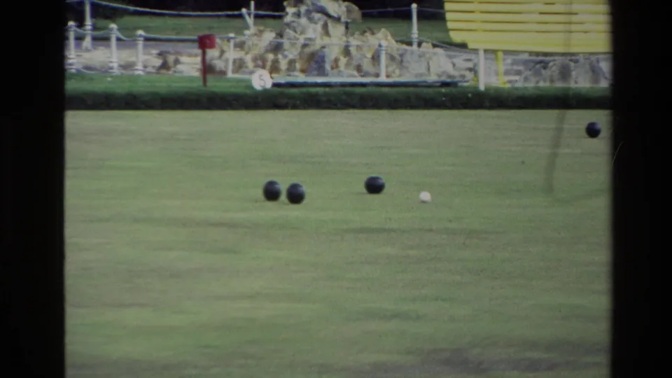 1981 New Zealand 8mm frame with bocce balls spread on a lawn court, a white pallino, stone edging, and a yellow bench in the background.