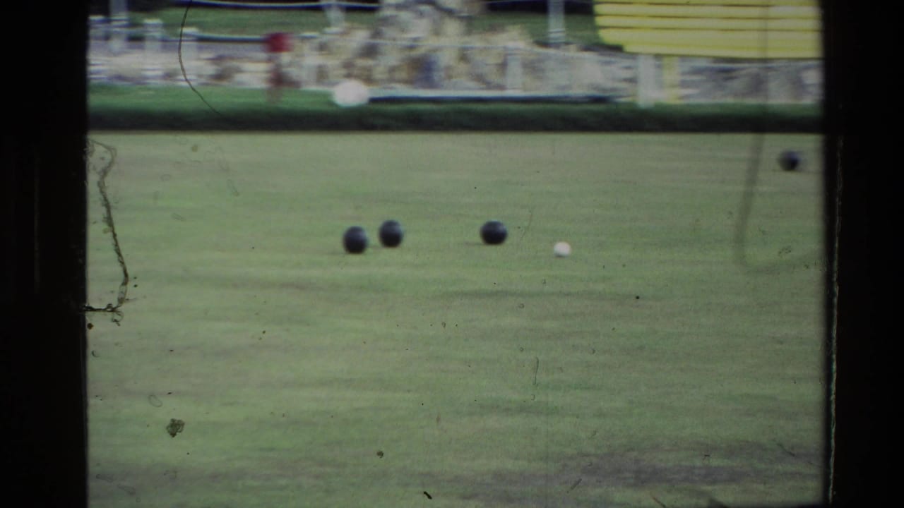1981 New Zealand home-movie frame showing a green lawn game area with dark balls, hedging, and a yellow building in the background.