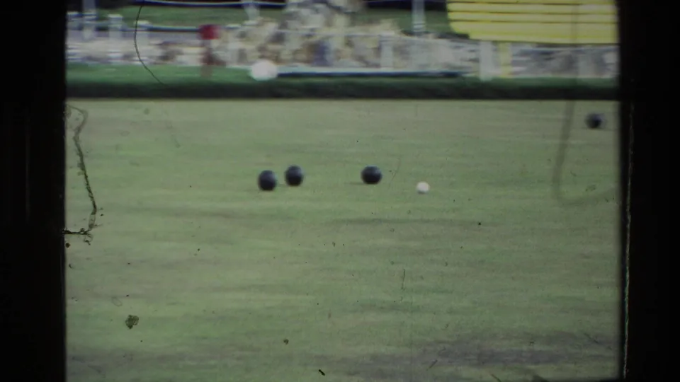 1981 New Zealand home-movie frame showing a green lawn game area with dark balls, hedging, and a yellow building in the background.