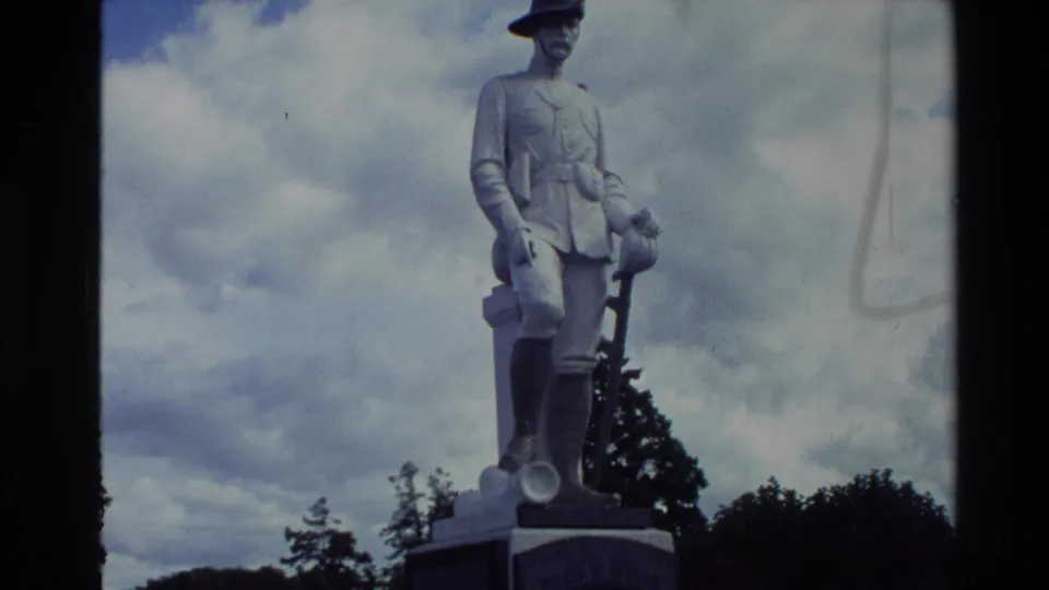 1981 New Zealand frame of a pale soldier monument on a pedestal, filmed from below against clouds and trees.