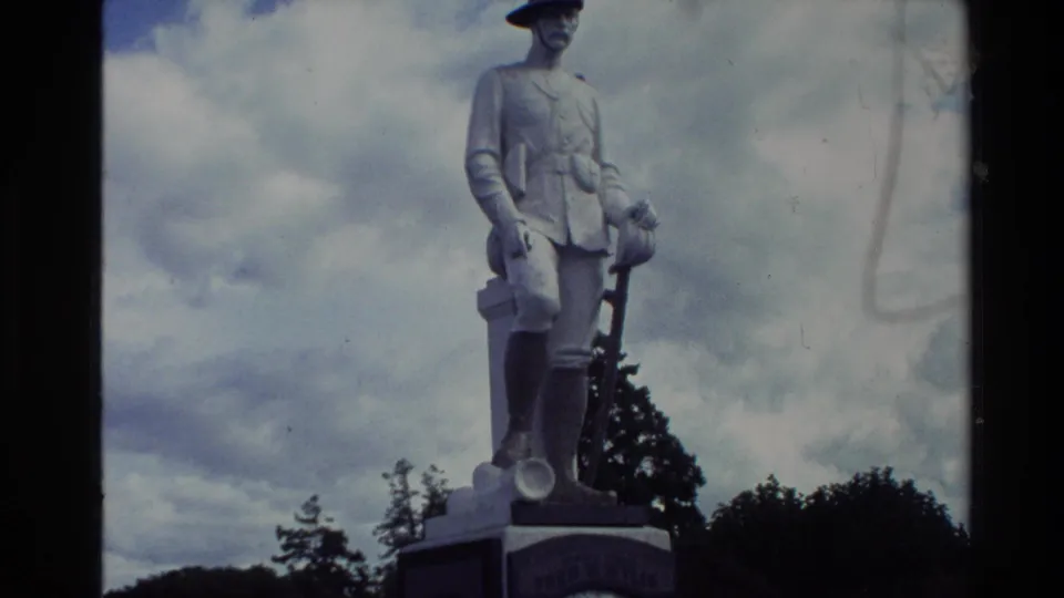 1981 New Zealand home-movie still holding on the white soldier memorial, with the pedestal partly visible below a clouded sky.