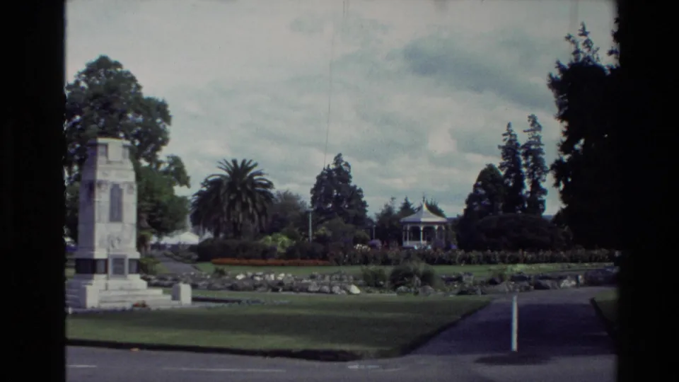1981 New Zealand wide view of a memorial garden with a monument, palms, flower beds, paved path, and small gazebo under overcast sky.