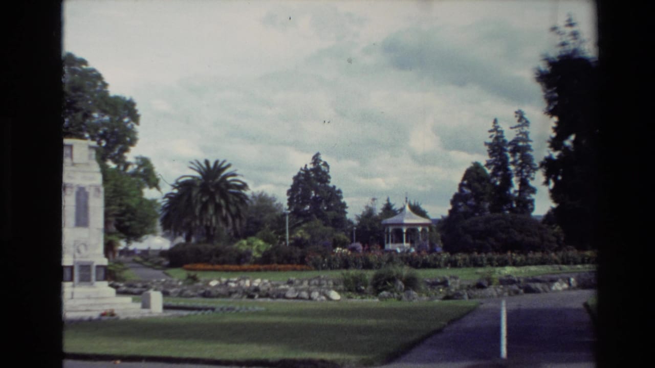 1981 New Zealand home-movie frame showing a formal park with lawns, a white memorial structure, flowerbeds, trees, and a distant bandstand.