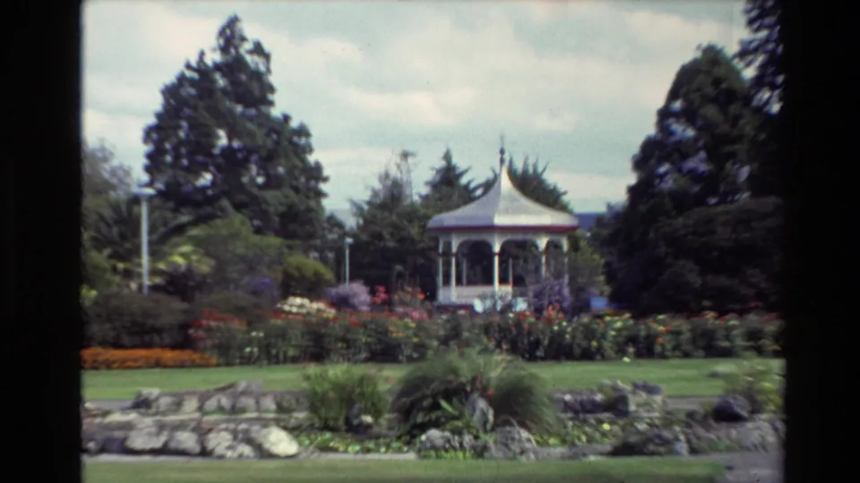 1981 New Zealand park scene centered on a white bandstand among flowerbeds, rocks, trees, lawns, and overcast sky.
