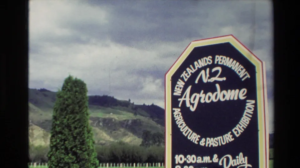 1981 New Zealand home-movie frame showing an Agrodome agriculture and pasture exhibition sign near rolling hills and a tall shrub.