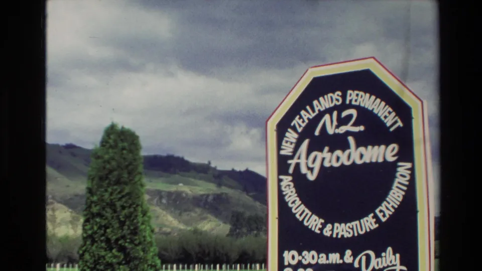 1981 New Zealand frame with the Agrodome sign, a tall shrub, rolling pastoral hills, and overcast sky.