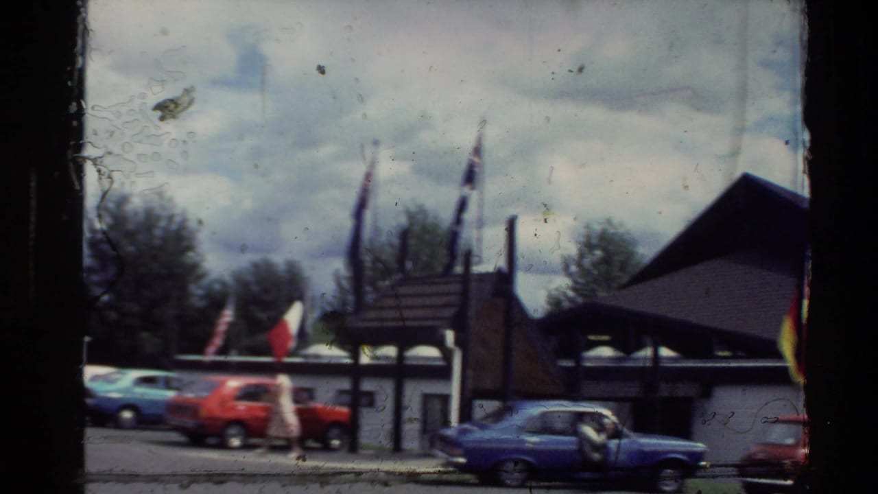 Exhibition building with international flags and parked compact cars under a cloudy sky.