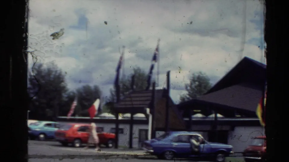 1981 New Zealand home-movie frame showing an exhibition building, flags, and parked blue and red compact cars.