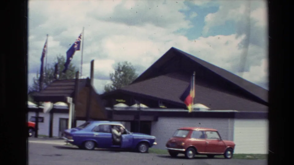 A blue sedan and red compact car sit outside the New Zealand exhibition building with flags visible above the entrance.