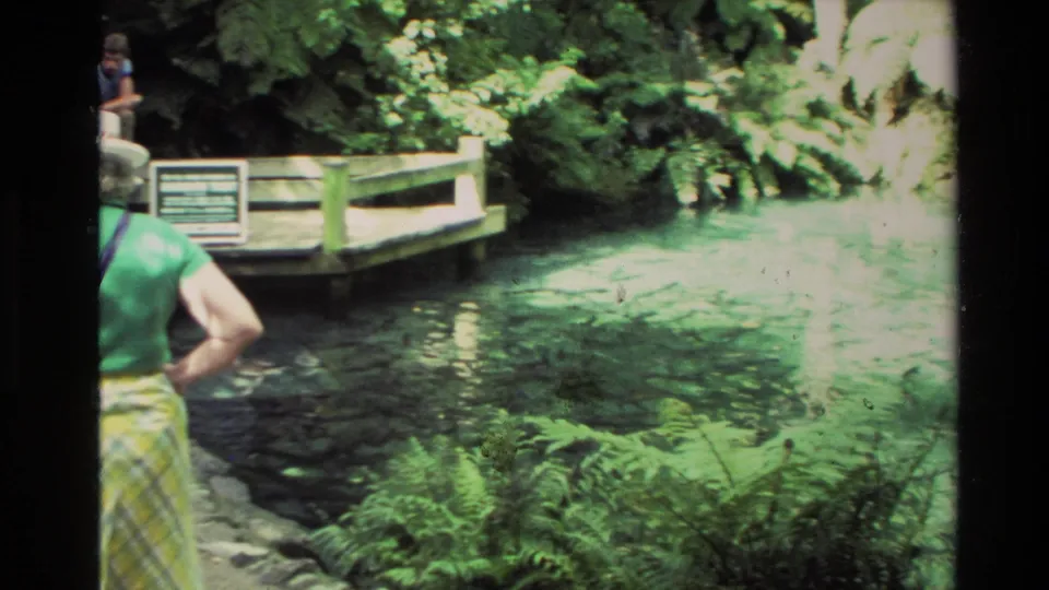 A woman in a green shirt and plaid skirt watches a turquoise pool from a dock area in 1981 New Zealand footage.