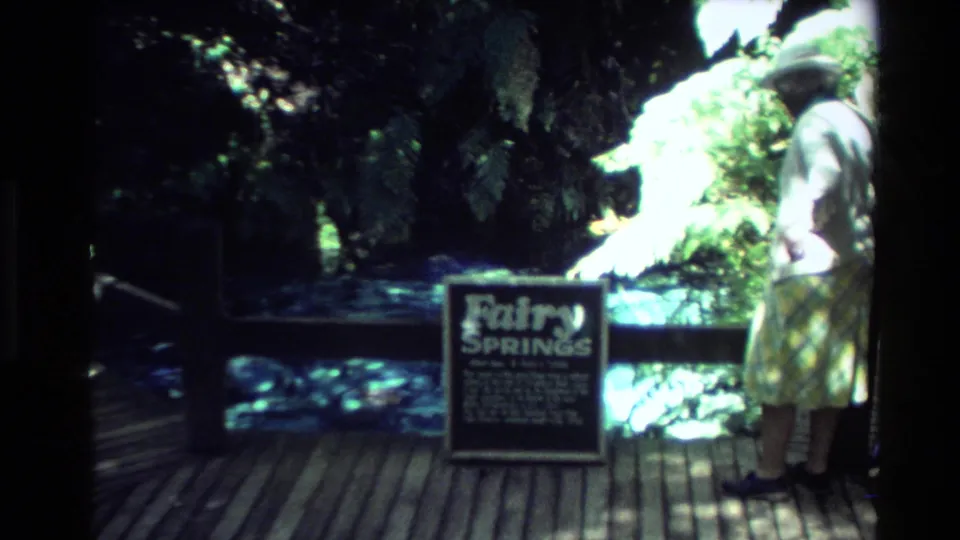 An adult woman in a light dress and hat stands beside a Fairy Springs sign on a shaded boardwalk in New Zealand footage.