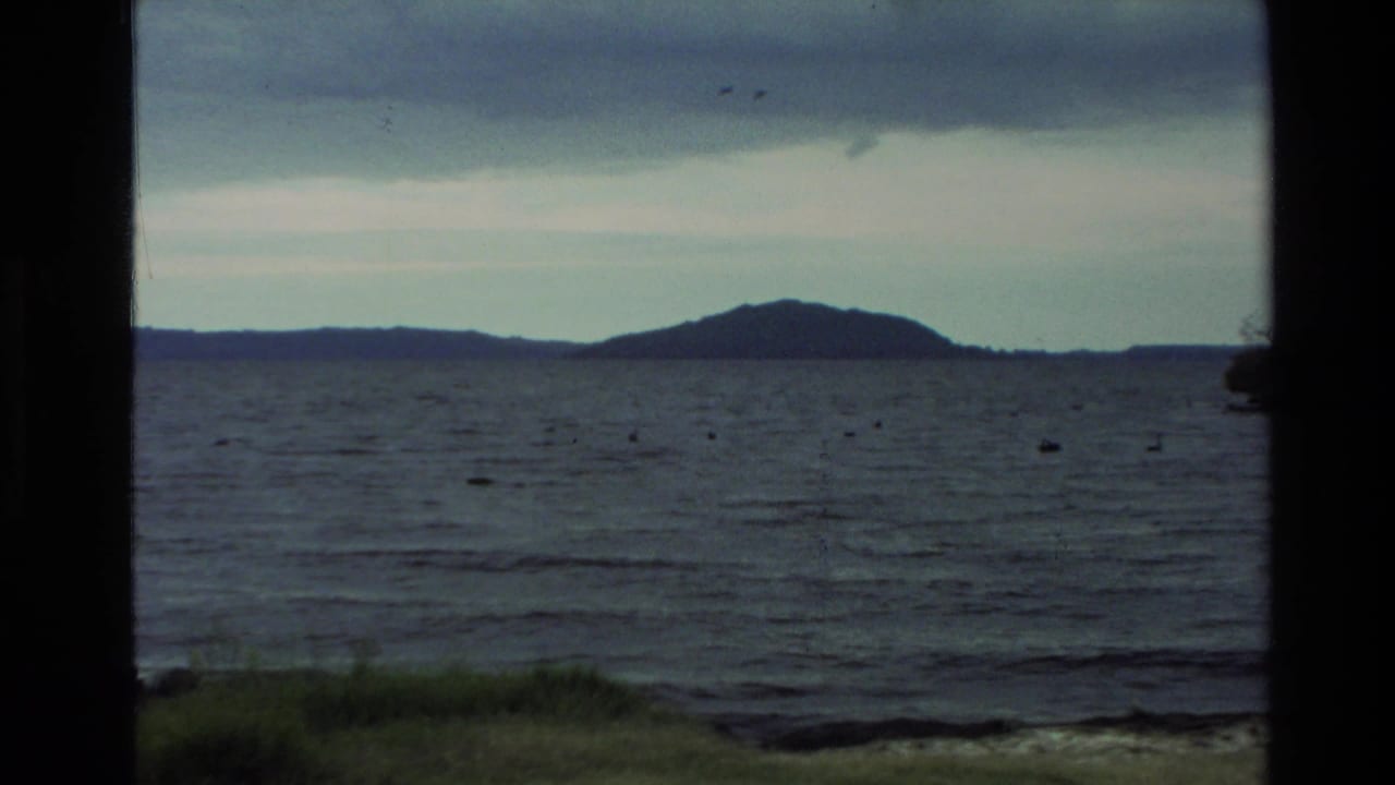 1981 New Zealand home-movie frame showing a calm lake, distant low hills, cloudy sky, and dark framing at the sides.