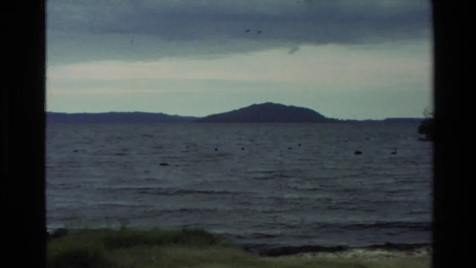 1981 New Zealand home-movie frame showing a calm lake, distant low hills, cloudy sky, and dark framing at the sides.