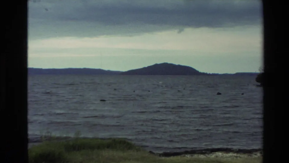 Wide 1981 New Zealand lake view with muted water, distant hills, cloudy sky, and dark window-like borders.