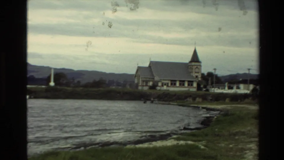 A shoreline church with a pointed steeple stands beside a New Zealand lake, with grassy banks and distant hills under cloud.