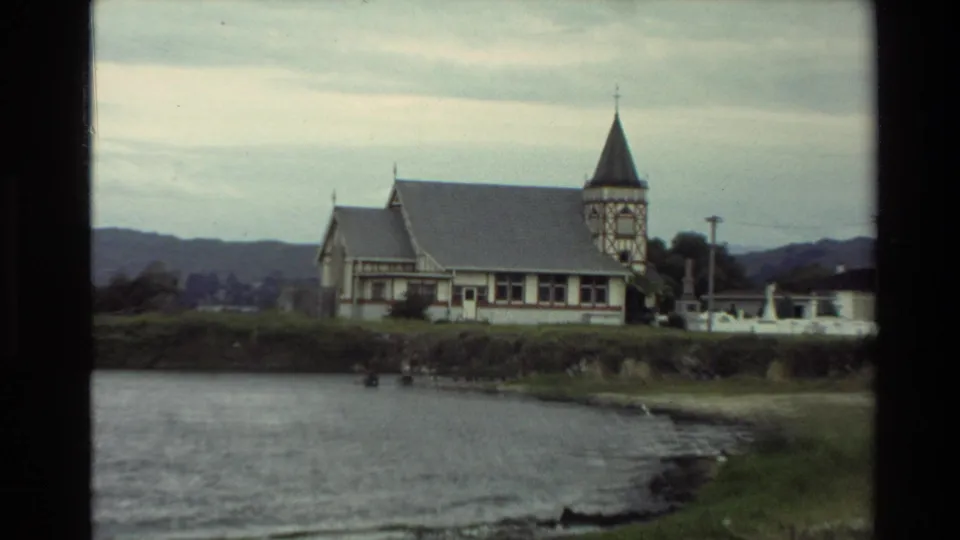 The 1981 New Zealand lakeside view shows a church near the shore, calm water in front, and low hills beyond.