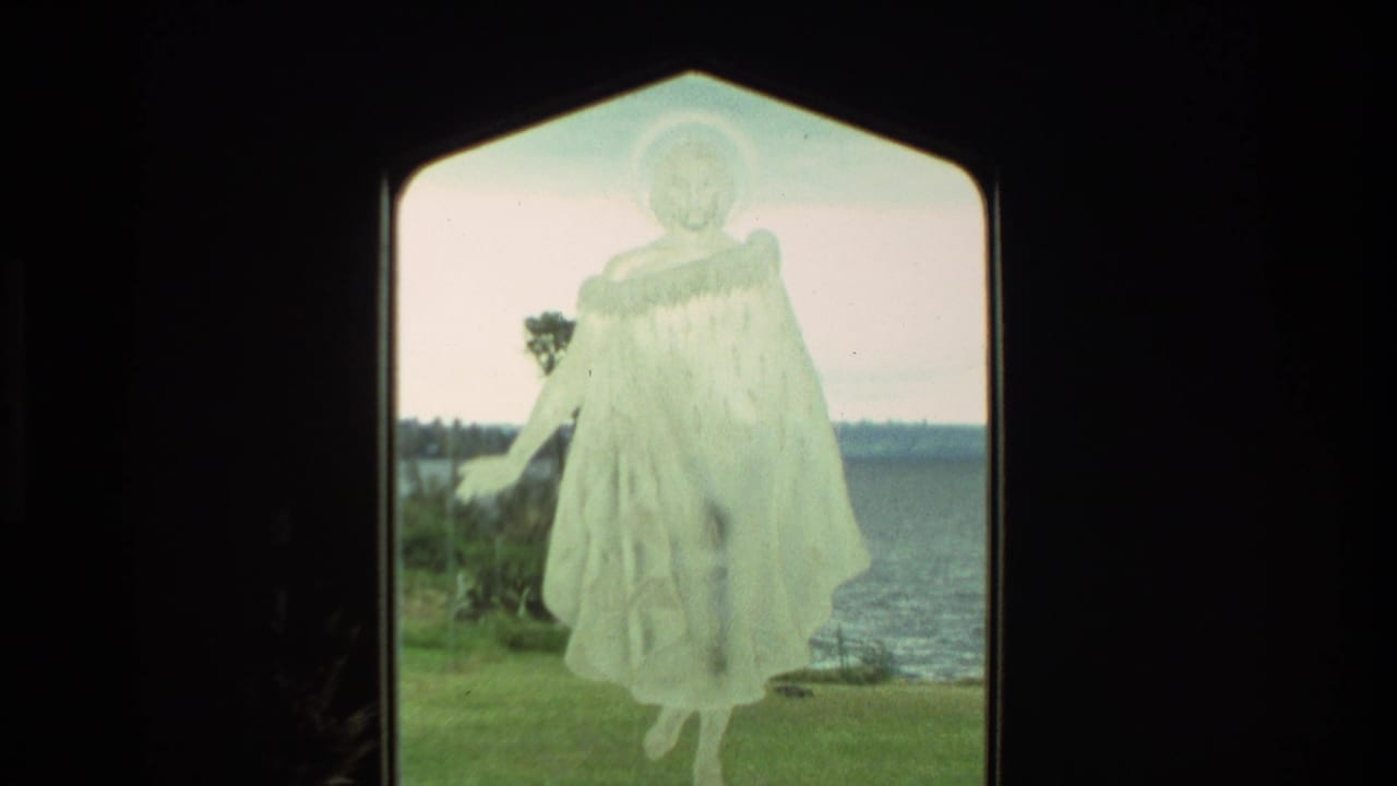 1981 New Zealand home-movie frame showing a pale robed statue or figure framed by a dark opening with grass and water beyond.