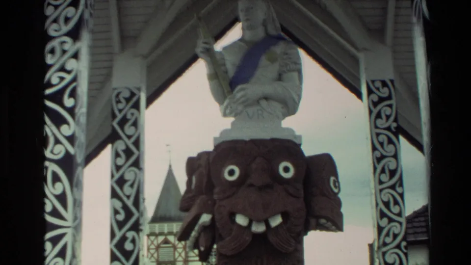 1981 New Zealand home-movie view of a carved figure and white statue inside an ornamented pavilion, with a pointed-roof building behind.