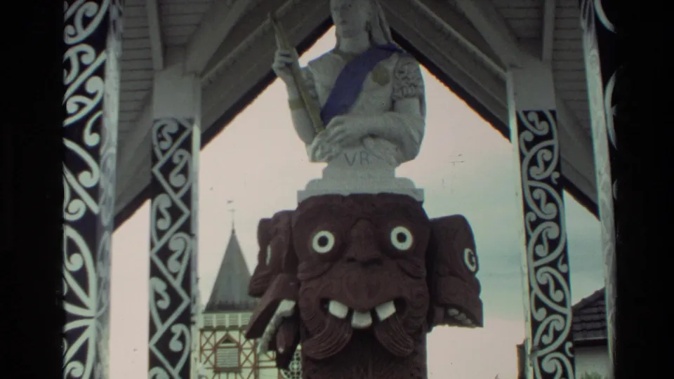 1981 New Zealand frame showing the carved face, partial white statue, blue sash, and ornamental black-and-white posts inside the pavilion.