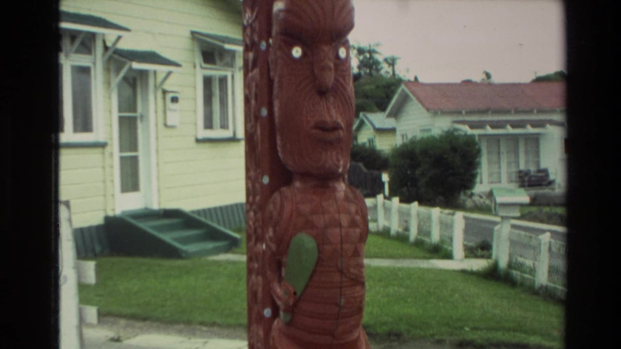1981 New Zealand home-movie frame showing a red-brown Maori carved post in a suburban street setting with yellow houses and lawns.