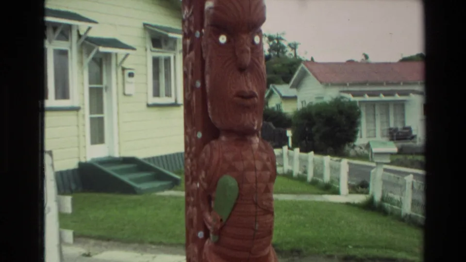 1981 New Zealand home-movie frame showing a red-brown Maori carved post in a suburban street setting with yellow houses and lawns.