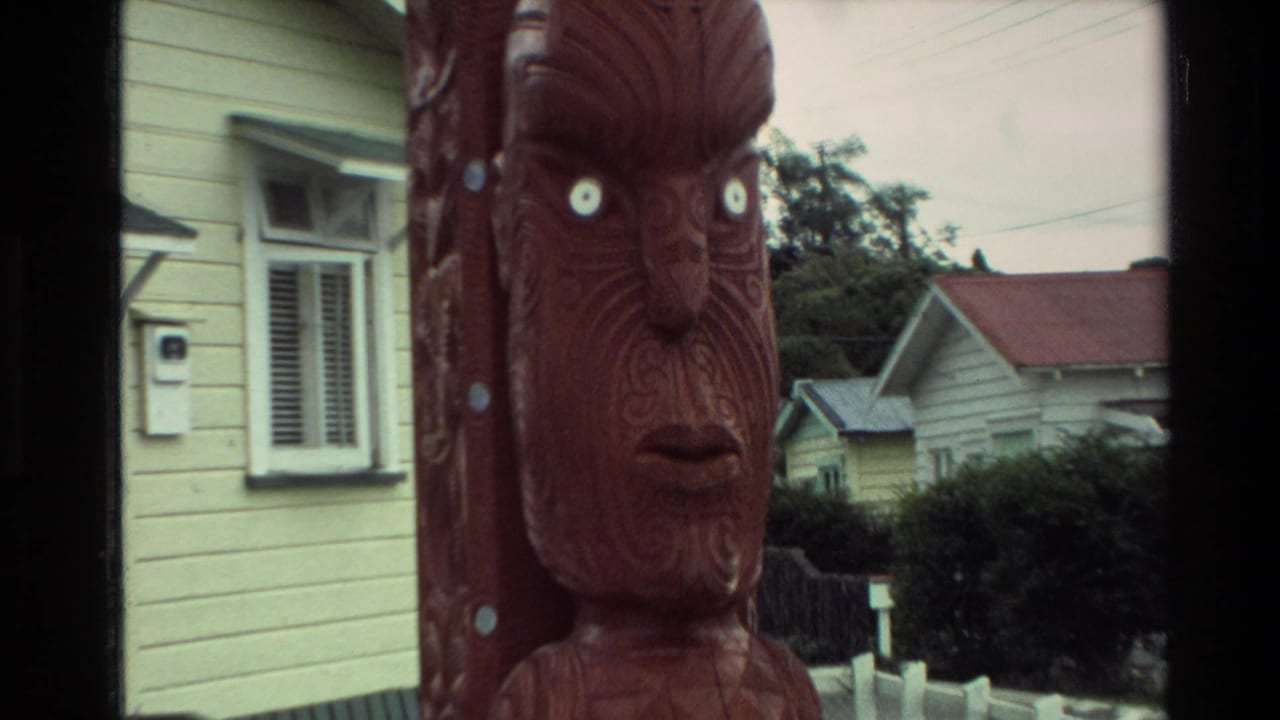 Close view of a carved wooden face beside a yellow house and distant roofs.