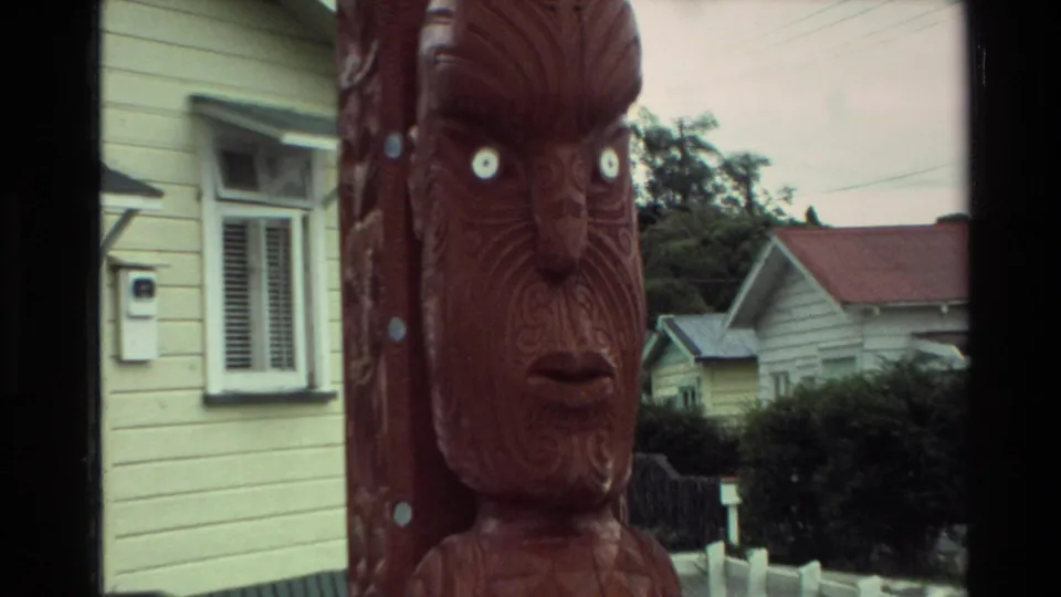 Close 1981 New Zealand frame of a Maori carved wooden face with white eyes, seen beside a yellow house and suburban roofs.