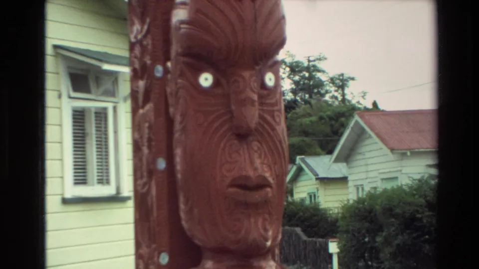 Tight home-movie view of a Maori carved wooden face with white eyes, framed against a yellow suburban house in New Zealand.