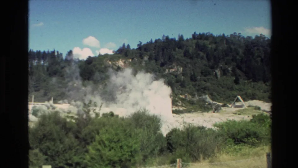 1981 New Zealand 8mm frame showing a wide visitor view of a geyser or thermal vent erupting with steam below wooded hills.