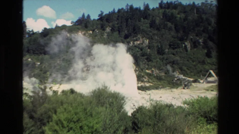 Wide 1981 New Zealand 8mm view of an active geyser throwing up steam against a green hillside and bright sky.