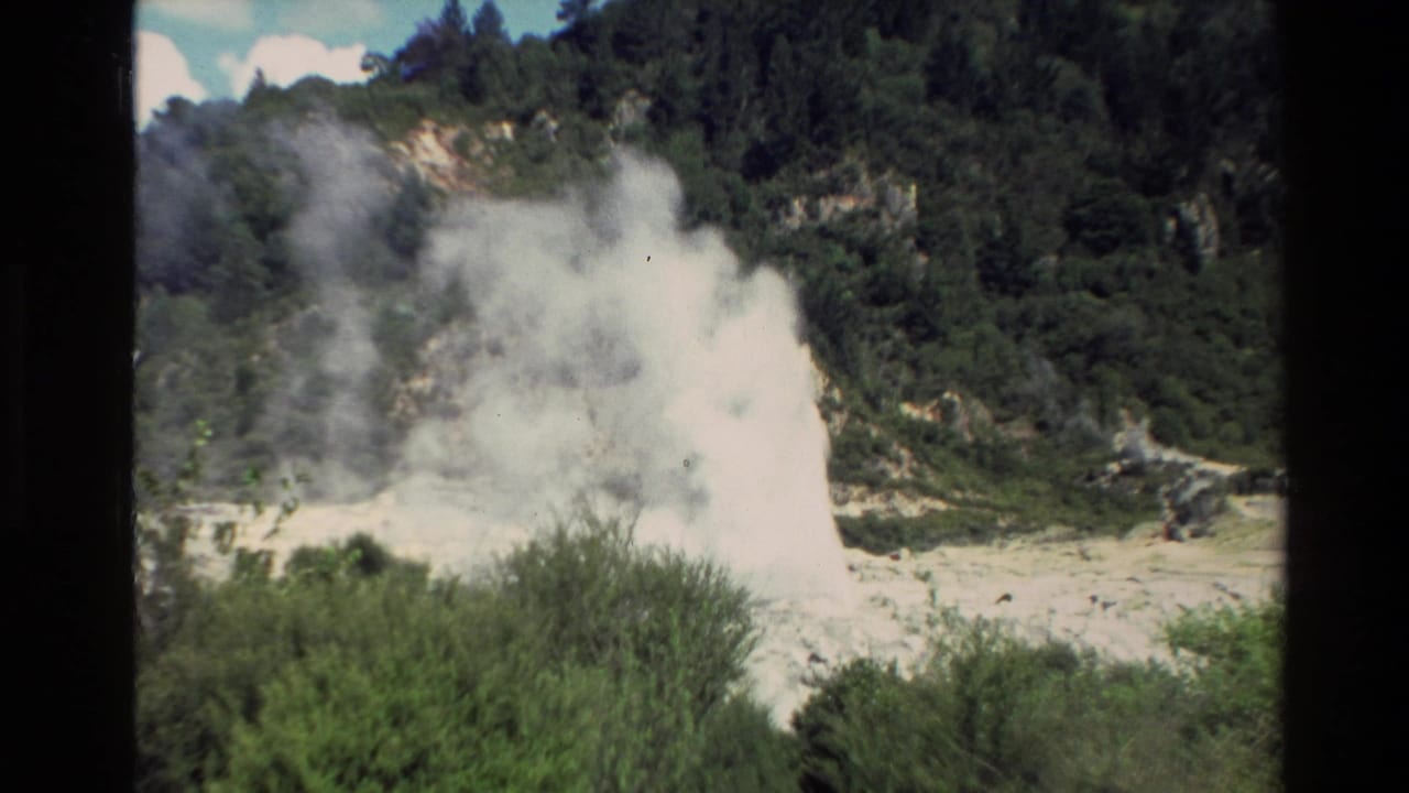 1981 New Zealand 8mm frame showing a geothermal geyser erupting white steam near shrubs below a wooded hillside.