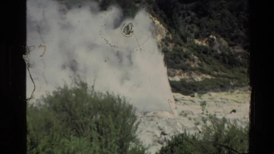 1981 New Zealand home-movie frame showing a large geyser steam cloud over shrubs, with film marks visible on the scan.