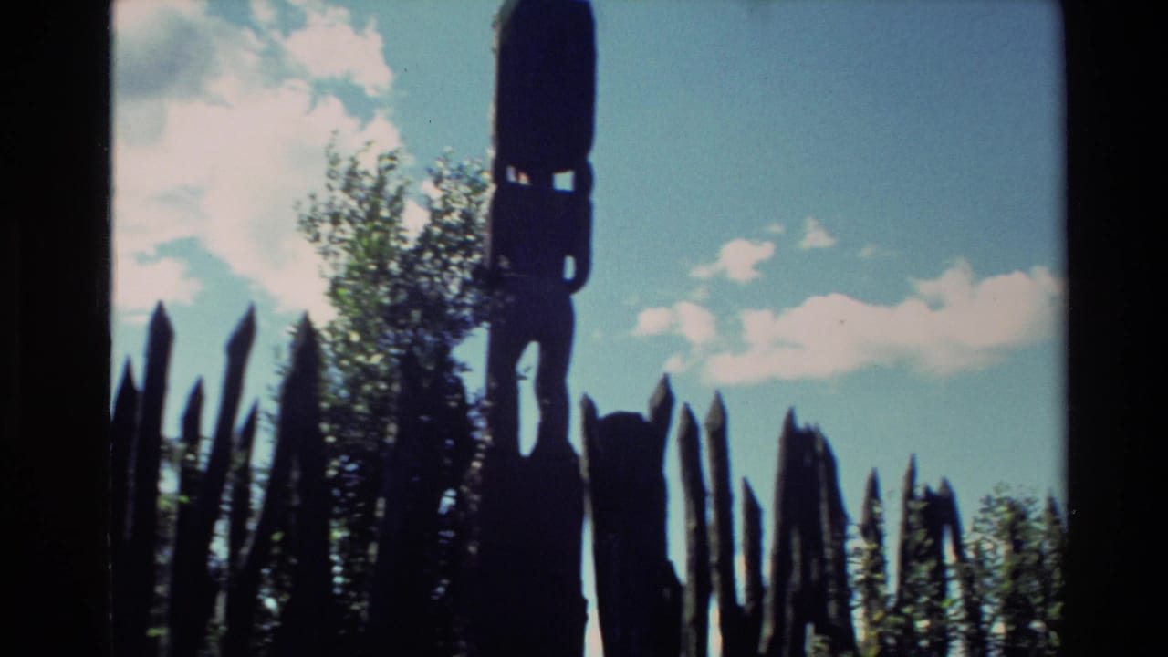 Dark carved figure and pointed wooden forms silhouetted against sky and trees.