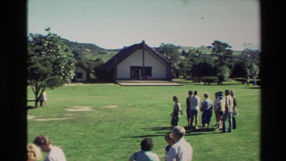 New Zealand 1981 8mm frame with visitors gathered on a lawn in front of a white gabled building, trees, and distant hills.