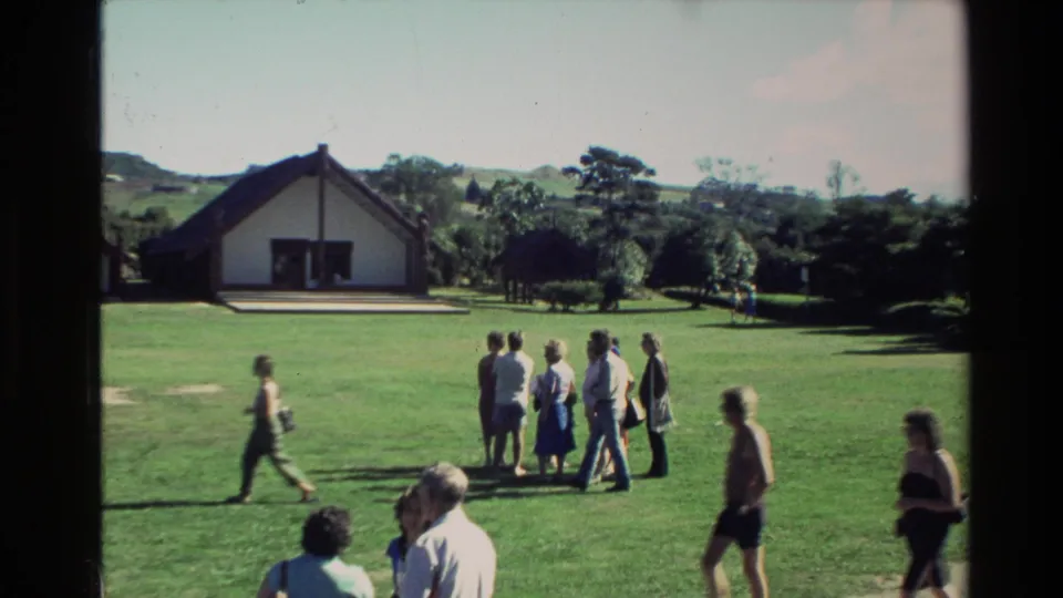 New Zealand 1981 8mm frame showing people walking and gathering on a green lawn with a white gabled building and hills behind.