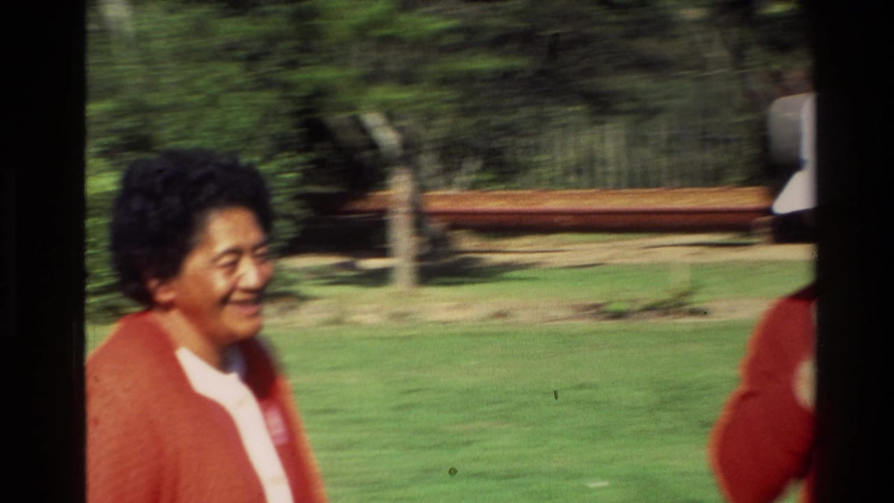 1981 New Zealand frame of a smiling woman in a red cardigan, with grass and trees blurred by handheld motion.