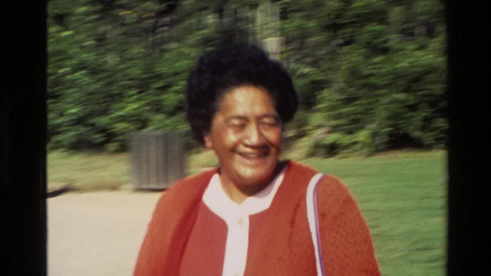 Close 8mm frame of a smiling woman in a red cardigan at the New Zealand outdoor site, with greenery behind her.