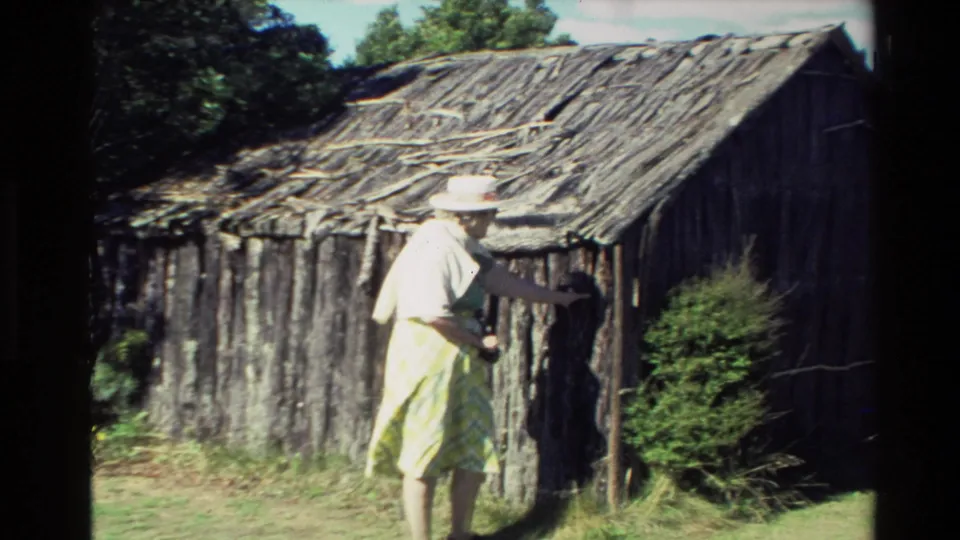 1981 New Zealand 8mm frame showing an adult woman in a brimmed hat and light clothing beside a rustic log-and-stick shack.