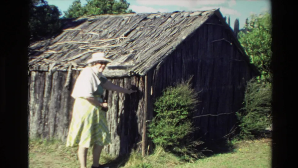 1981 New Zealand frame with an adult woman inspecting a weathered stick-and-log shack under bright sun, with shrubs and trees around it.
