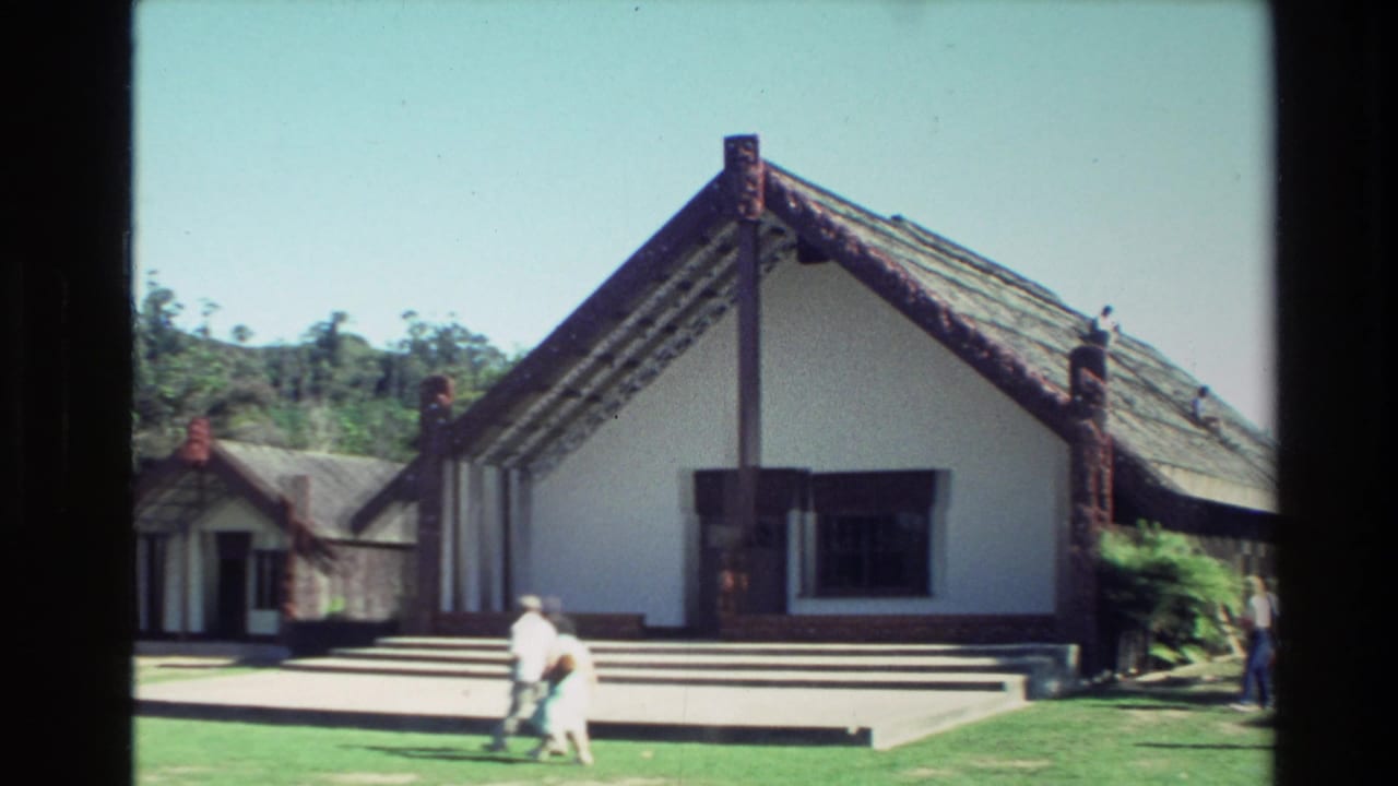 1981 New Zealand home-movie frame showing a Maori meeting house exterior with carved posts, roof detail, lawn, and visitors passing by.