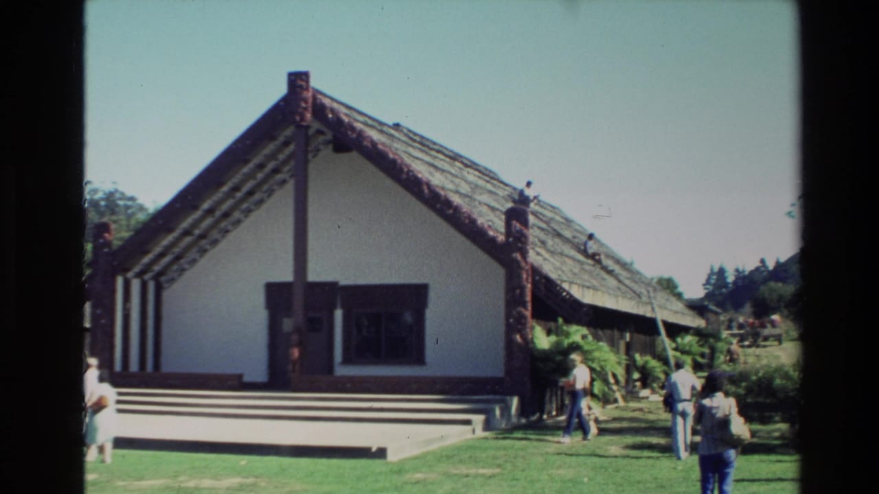 1981 New Zealand frame of a traditional Maori meeting house exterior, with people strolling on the lawn under a clear sky.