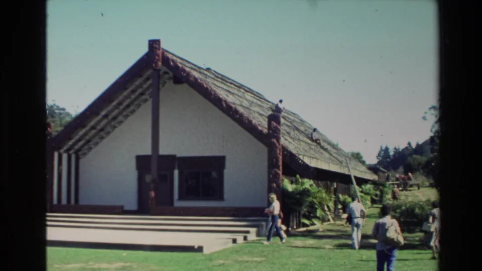 Angled 1981 New Zealand home-movie view of a Maori meeting house with carved posts, steps, lawn, trees, and visitors in motion.